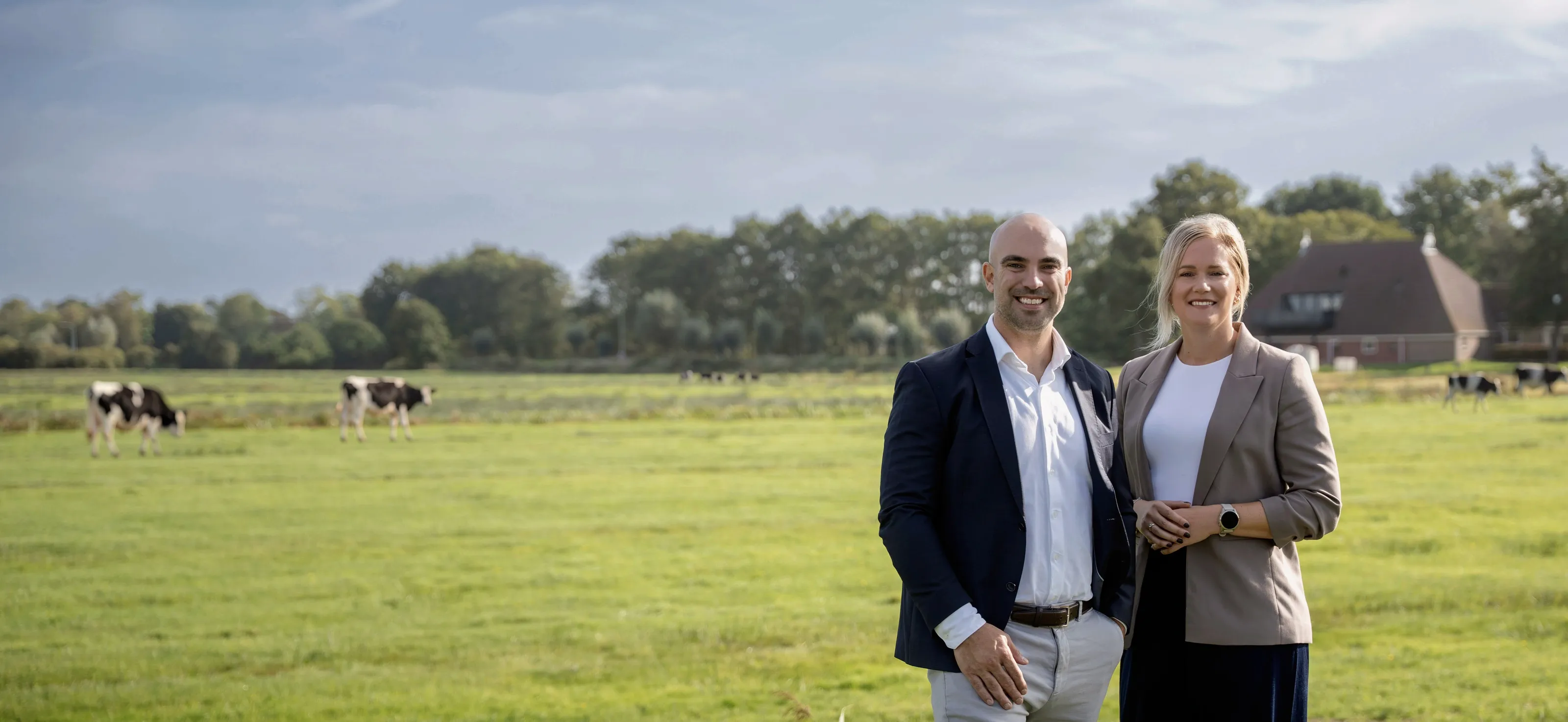 Tim de Vries and Elina van der Schaaf standing in a field with grazing cows, a farmhouse and a row of trees behind them